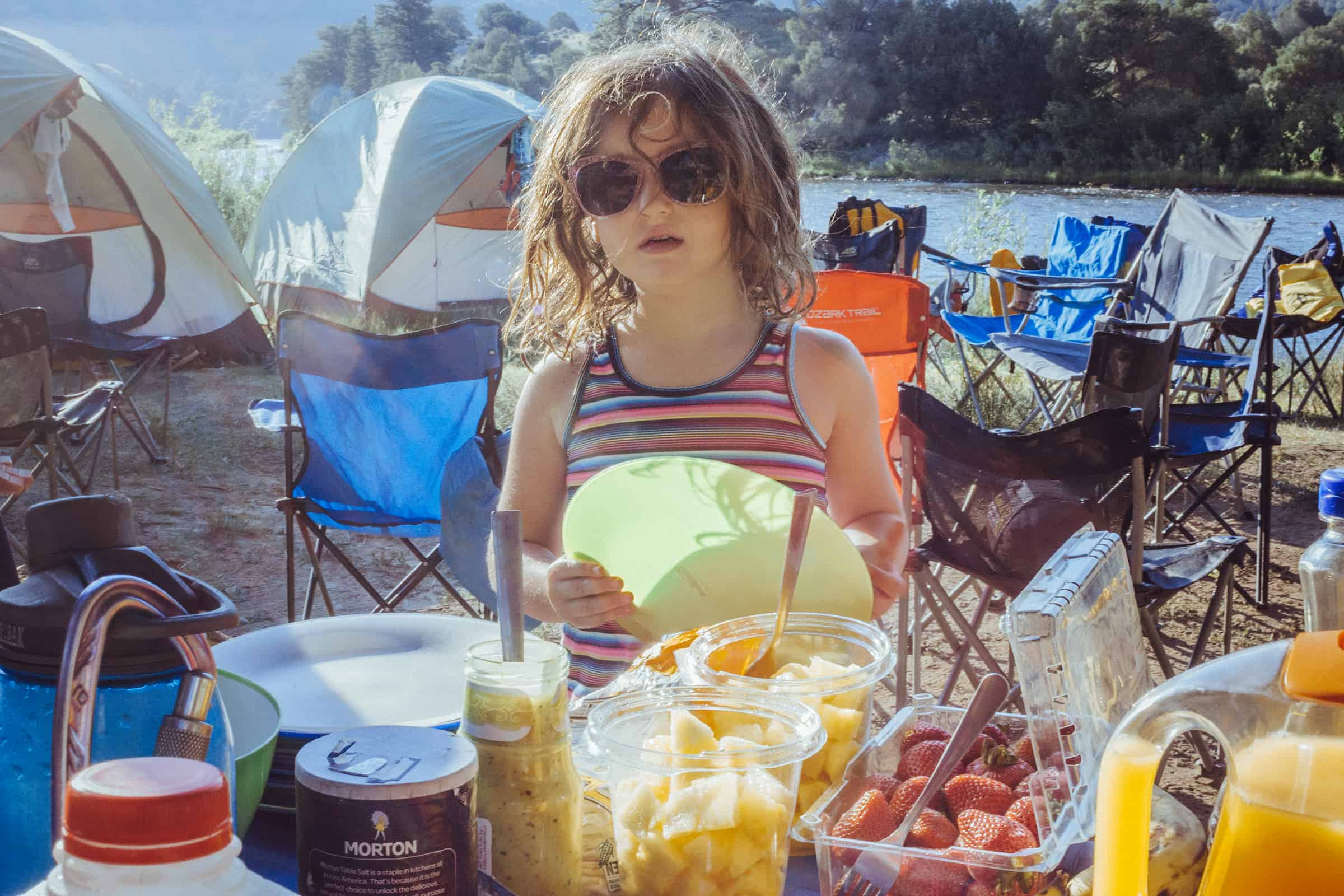 Child looks for a breakfast serving by a table filled with food on the river bank with camping chairs and tents in the background